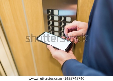 Man hands holding blank screen mobile phone next to elevator control panel.