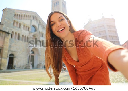 Smiling tourist girl taking selfie photo in Parma City, Italy. Female traveler take self portrait in Piazza Duomo square in Parma, the Italian Capital of Culture 2021.