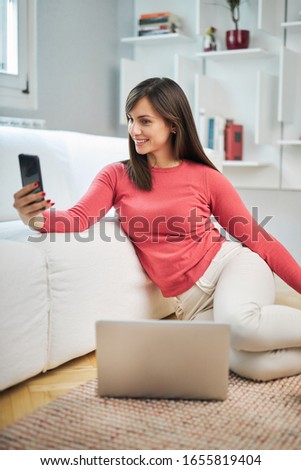 Young attractive smiling woman sitting on the floor at home, taking selfie and relaxing.