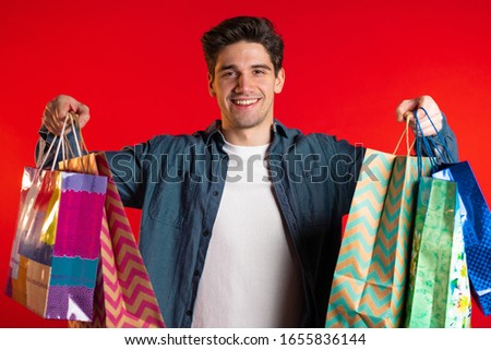 Handsome young man holds shopping paper bags on red studio background. Guy bought presents on sales with discounts.