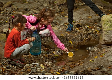 Children pump water into a plastic bottle. Girl draws water from a mountain stream. Friends replenish their drinking water. Dirty spring with turbid water. A hike with survival skills.