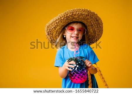 little blonde girl in a straw hat and sunglasses is smiling on a yellow background. child girl in a blue dress is holding a pineapple-shaped drink glass on a yellow background.summer vacation concept
