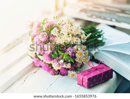 A bouquet of wild flowers, a book and a small gift on the wooden windowsill, selective focus.