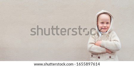 Little boy 3-5 years old, beige wall background, emotions joy, fun, laugh, laughter, pleasure happiness. Knitted warm sweater with hood. Free space for copy text. Holiday concept banner for print