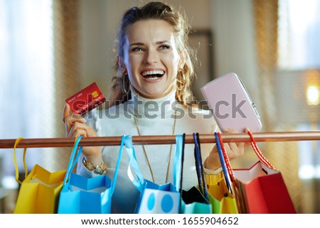 happy stylish female in white sweater and skirt with credit card and wallet near colorful shopping bags hanging on copper clothes rail.
