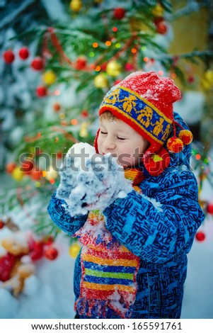 A laughing boy in a red patterned knitted cap with ear-flaps and with a bright scarf playing with snow near the decorated Christmas tree on a frosty winter day outdoor