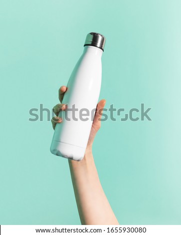Close-up of female hand holding white reusable steel stainless thermo water bottle isolated on background of cyan, aqua menthe color. Plastic free. Royalty-Free Stock Photo #1655930080