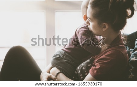 Concerned woman holding and hugging a small child. Mom from the generation of milenials sits by the window and holds the son. Slight noise and shallow depth of field.