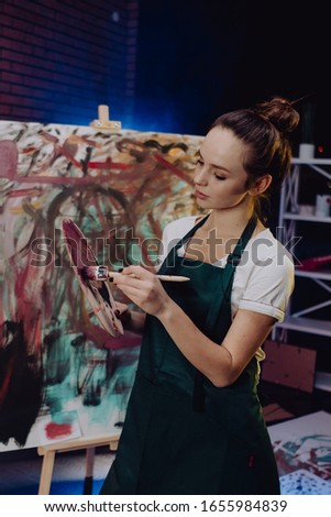 A young female artist is creating an abstract painting. She is working in the loft space with dimmed lights. She is using oil paint and a brush.