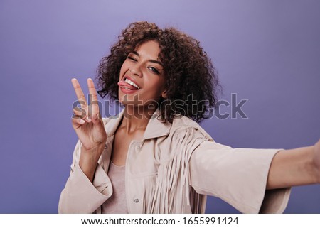 Brunette girl in beige outfit winks, takes selfie, shows tongue and peace sign. Portrait of positive woman in jacket smiling on purple backdrop