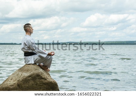 A man in a kimono sits on a rock, under it is the sea.  Master with a black belt.  Practical class in karate.  The technique of meditation.  Cleansing and thinking about life before the fight.  Royalty-Free Stock Photo #1656022321
