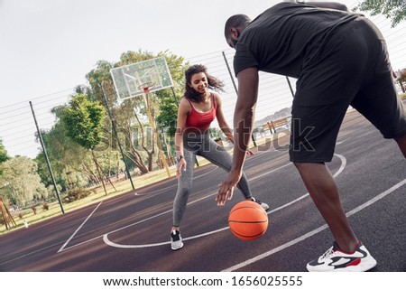 Young african descent couple man dribbling back view close-up while woman standing in front blocking smiling concentrated following ball on basketball court outdoors