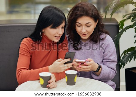 Photo of two busy curious good looking young females using their smartphones, chatting, sitting at table in cafeteria, drinking coffee, looking at screen attentively. People and net addiction concept.