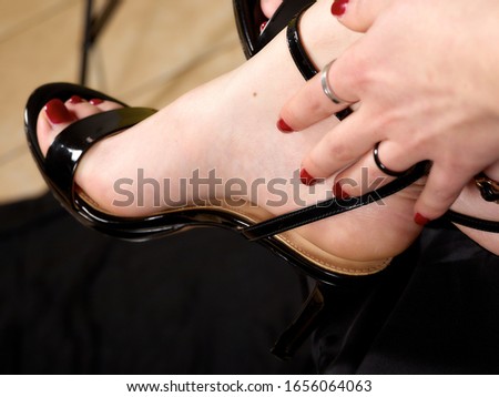 Woman foot in black patent leather sandals and high heel with woman hand on black satin black background.