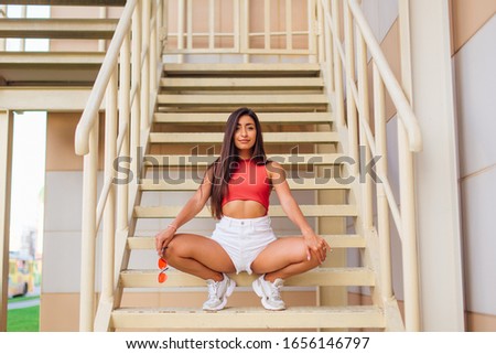 Street fashion summer portrait of a woman sitting on stairs holding pink sunglasses in hand outdoors. Full-length portrait young beautiful model in shorts posing on the stairs. Copy space