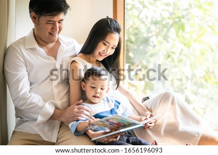 Family happy activity. Asian mother holding book to their daughters sitting on sofa at home. Little cute kid writing on the book drawing a picture and Older sister and beautiful mom looking on it.
