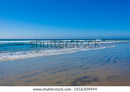 A cloudless day on a beach in California with foamy blue and teal waves cascading on wet sand
