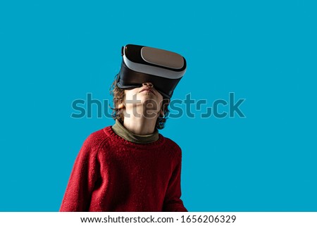 7 years old boy wearing vr goglles playing games or watching a 3d video.  Kid watching virtual reality 360 degree video on a flat blue background