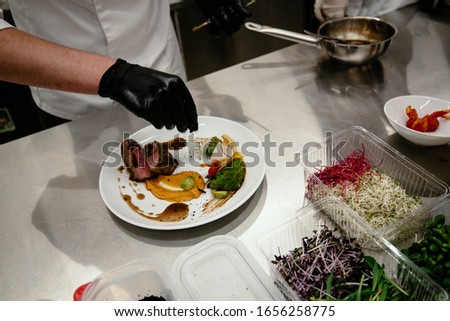 Chef plating up food in a restaurant pouring a gravy or sauce over the meat before serving it to the customer. Appetizing steak with vegetables on a white plate. Exquisite restaurant food