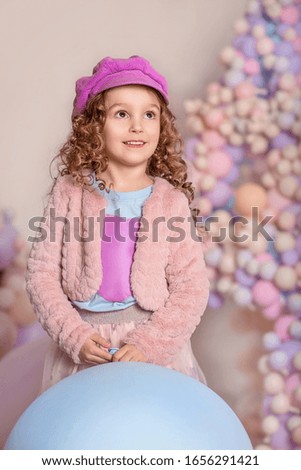 Emotional curly-haired girl in a purple hat among colorful balloons. Pastel color.