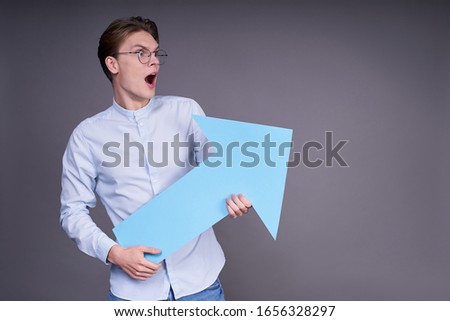 Cute cheerful charming young Caucasian guy, 20 years old, in a blue shirt, holding a big blue arrow in his hands, shocked, indicates the direction to the right, posing on a gray background.