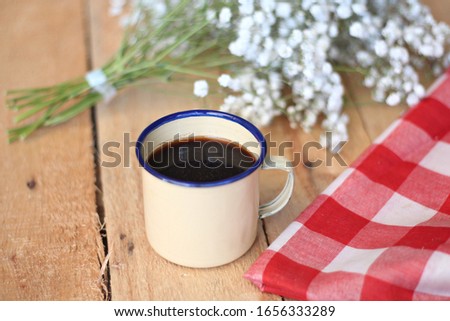 
a traditional cup of tin with coffee in it on a wooden table with baby breath flowers in the background and a checkered tablecloth beside it.