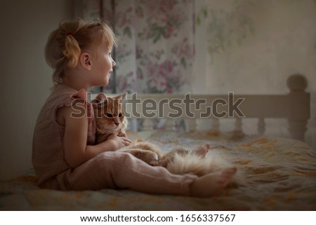 A pretty little girl with tails sits on the bed and hugs a red cat. Love of children and animals. Taking care of animals.