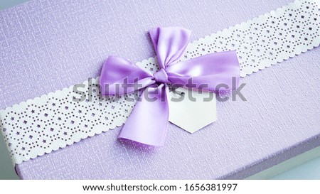 Top view of a lilac gift box with through patterns on a white ribbon, a purple bow and a label for inscription. The concept of a holiday, congratulations and surprises.
