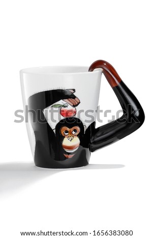 Subject shot of a ceramic mug with a painted monkey with an apple, a handle in the form of the monkey's arm and a white inner surface. The cute cup is isolated on the white background with shadows. 