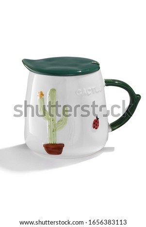 Subject shot of a white ceramic mug with a picture of a flowered cactus in a pot and a ladybird, a figured handle and a green leaf-formed cover. The cute cup is isolated on the white background. 