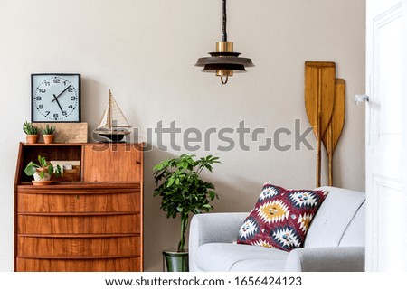 Stylish retro composition of living room interior with vintage wooden cabinet, gray sofa, plants, pillow, clock, paddle, pendant lamp and elegant personal accessories. Retro home decor Template. 