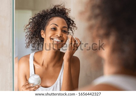 Black girl take care of her beautiful skin. Young african woman applying moisturizer on her face. Smiling black natural girl holding little jar of skin lotion in bathroom for beauty treatment routine. Royalty-Free Stock Photo #1656442327