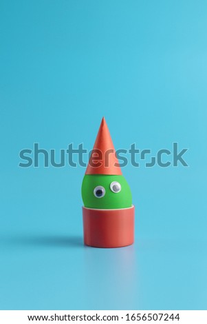 One green egg with eyes in a red festive cap in a round red box on a blue background. Minimal Happy Easter concept decoration. Copy space for text mockup. Close up photography. 