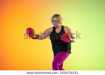 Young caucasian plus size female model's training on gradient orange background in neon light. Doing workout exercises with boxing gloves. Concept of sport, healthy lifestyle, body positive, equality.