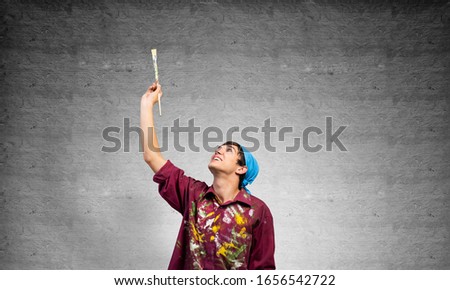 Young painter artist pointing upwards. Man holds paintbrush over his head. Portrait of caucasian painter on gray wall background. Creative hobby and artistic occupation. Art school student posing.