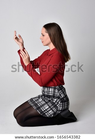 full length portrait of a pretty brunette girl wearing a red shirt and plaid skirt with leggings and boots. Seated pose on a  studio background.
