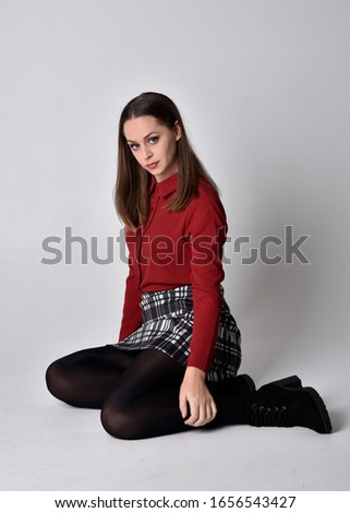 full length portrait of a pretty brunette girl wearing a red shirt and plaid skirt with leggings and boots. Seated pose on a  studio background.
