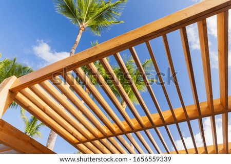 Wooden sunshade roof structure fragment under blue sky at sunny summer day, palm trees are on a background