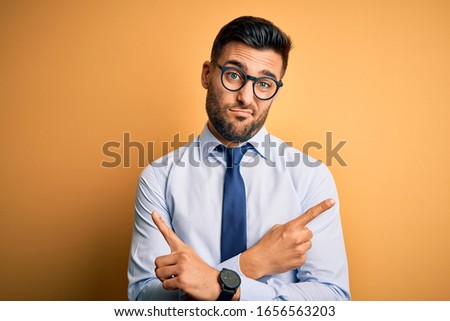 Young handsome businessman wearing tie and glasses standing over yellow background Pointing to both sides with fingers, different direction disagree