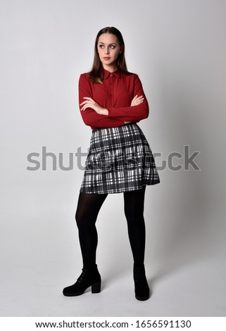 full length portrait of a pretty brunette girl wearing a red shirt and plaid skirt with leggings and boots. Standing pose on a  studio background.
