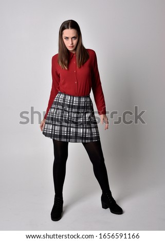 full length portrait of a pretty brunette girl wearing a red shirt and plaid skirt with leggings and boots. Standing pose on a  studio background.
