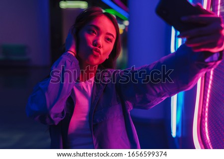Beautiful young asian woman with long dark hair standing infront of a neon wall, taking a selfie