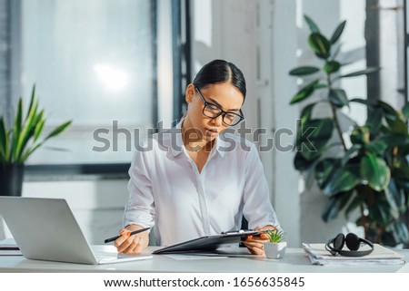 asian translator in eyeglasses working online with laptop and documents in office