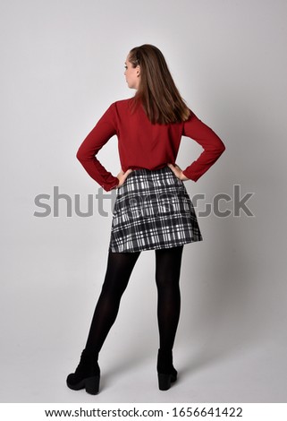 full length portrait of a pretty brunette girl wearing a red shirt and plaid skirt with leggings and boots. Standing pose facing away from the camera on a  studio background.