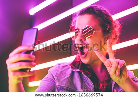 Portrait of modern young girl in round glasses making selfie at the neon light. Shows sign pis.