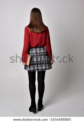 full length portrait of a pretty brunette girl wearing a red shirt and plaid skirt with leggings and boots. Standing pose holding a book, on a  studio background.