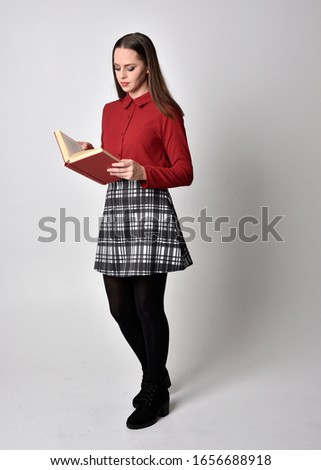 full length portrait of a pretty brunette girl wearing a red shirt and plaid skirt with leggings and boots. Standing pose holding a book, on a  studio background.