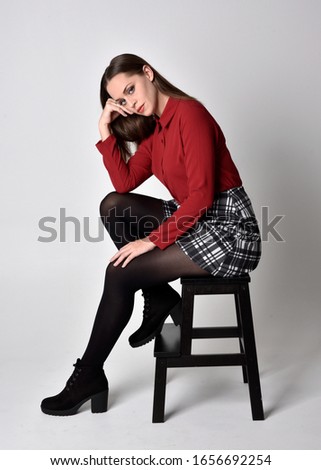 full length portrait of a pretty brunette girl wearing a red shirt and plaid skirt with leggings and boots. Sitting on a chair against a  studio background.