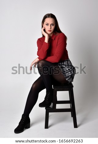 full length portrait of a pretty brunette girl wearing a red shirt and plaid skirt with leggings and boots. Sitting on a chair against a  studio background.