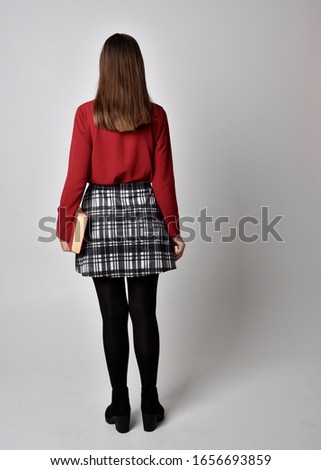 full length portrait of a pretty brunette girl wearing a red shirt and plaid skirt with leggings and boots. Standing pose, holding books, against a  studio background.